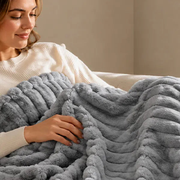 A woman reclining on a bed or sofa, covered with a plush, ribbed gray throw blanket. She is wearing a cream-colored knit sweater, and the background is a soft beige wall.