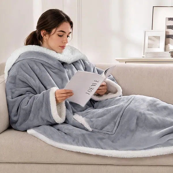 A woman sits on a beige sofa reading a book while wearing a large light gray wearable heated blanket with sleeves. The blanket covers her body and lap, and the room is softly lit with a neutral interior background.